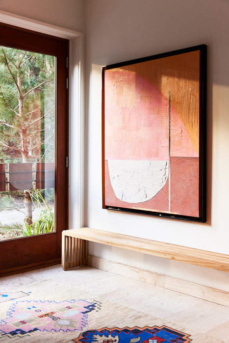 abstract painting in pink hues with brown acrylic frame hanging above a slatted wooden bench in hallway