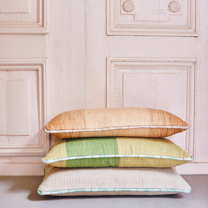 3 large hand woven woolen lumbar pillows with green and white piping on a stack against a wooden wall