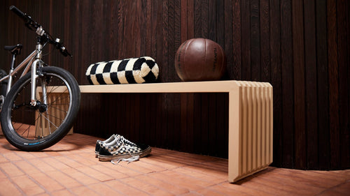 sand colored wooden bench against a dark brown wall with a checkered bolster pillow and a bike, sneakers and basketball