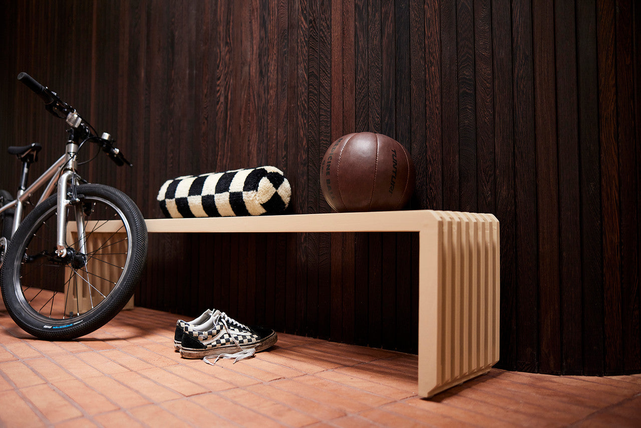 sand colored wooden bench against a dark brown wall with a checkered bolster pillow and a bike, sneakers and basketball