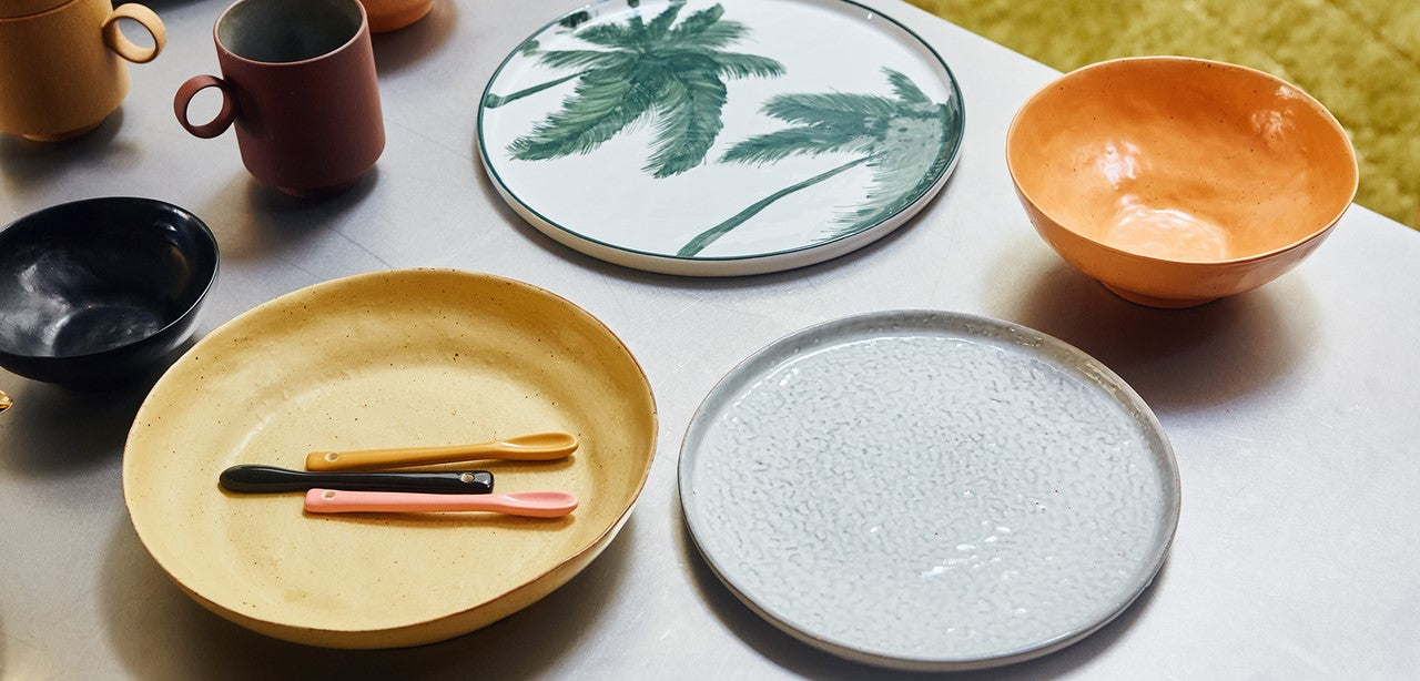 deep yellow plate with stoneware teaspoons in 4 colors and a white large basic dinner plate next to an orange bowl and a plate with green palm trees