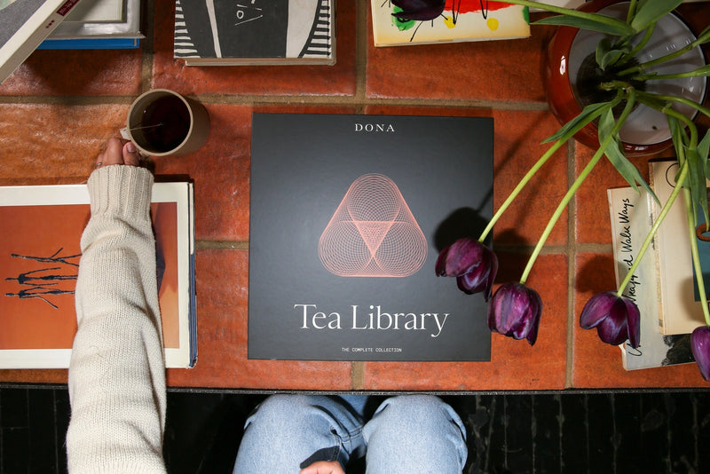Person holding a cup of tea next to a 'Tea Library' box on a wooden table with tulips.