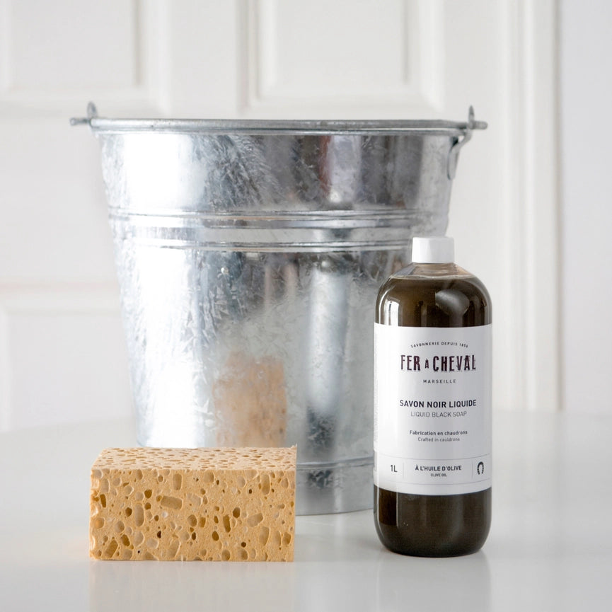 Bottle of 'Fer a Cheval' soap, sponge, and metal bucket on a white background