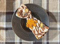 Decorative napkin with floral pattern and orange tassels on a dark plate against a checkered tablecloth.