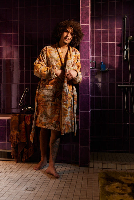 man in bathroom with purple tiles wearing a bathrobe with pockets and orange brown palm tree motive