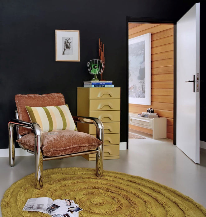 room with black wall, chrome and brown chair chest of drawers in yellow and a large round rug with polaroid's and magazine 