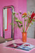 tall long standing chrome chubby mirror on pink wall and orange vase with flowers