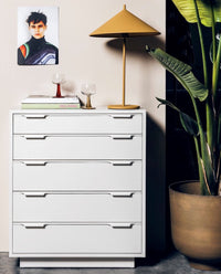 white wooden dresser with 5 drawers with yellow tablelamp and metal planter with plant