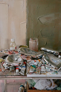 Art supplies including paint tubes, brushes, and jars on a table with a textured wall background.