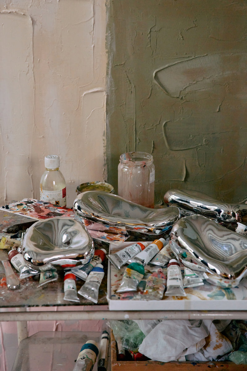 Art supplies including paint tubes, brushes, and jars on a table with a textured wall background.