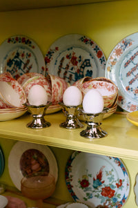 silver egg cups whit white eggs in a yellow cabinet with flower plates