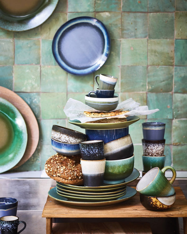 Collection of ceramic plates and mugs on a wooden table against a green tiled wall.