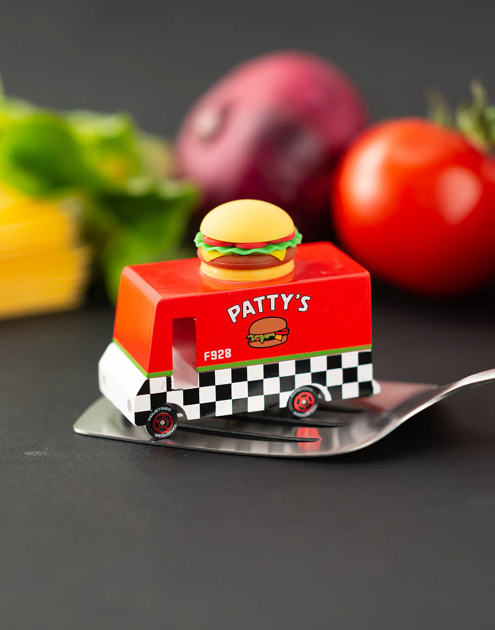 Toy truck with a hamburger on top, resembling a cheeseburger, on a dark surface with vegetables in the background.