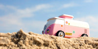 Pink and white vintage-style camper van on sandy terrain with a blue sky.