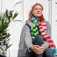 Woman wearing colorful striped scarves sitting against a white wooden wall.
