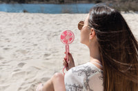 Woman sitting on a beach holding a pink handheld fan.