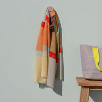 Colorful scarf draped over a chair with a woven bag on a wooden stool against a light gray background