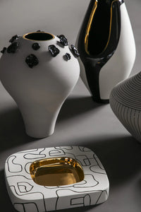 Decorative ceramic items including a white vase with black accents, a black and gold pitcher, and a square dish with gold interior on a gray background.
