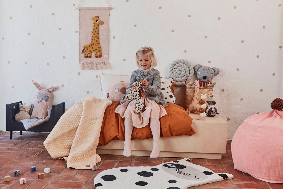 Child sitting on a bench with stuffed animals in a room with polka dot walls and toys on the floor.