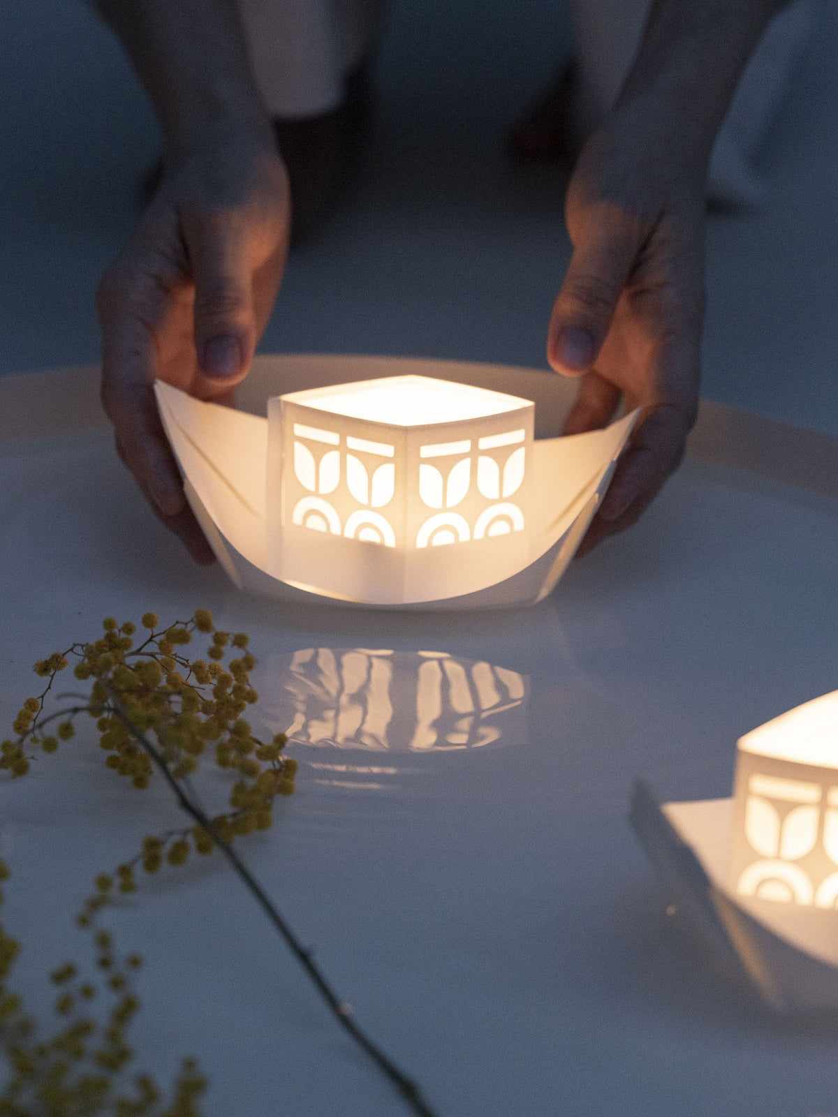 Hands holding a lit paper boat with another boat in the background on a reflective surface.