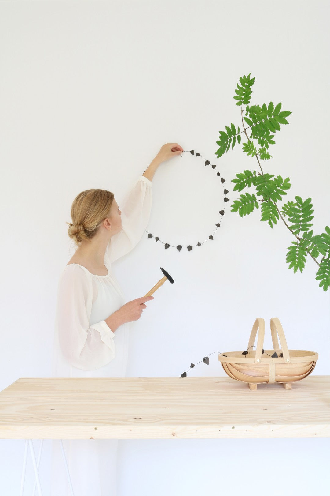 Person hanging a plant on a white wall with a wooden table and basket in the foreground.