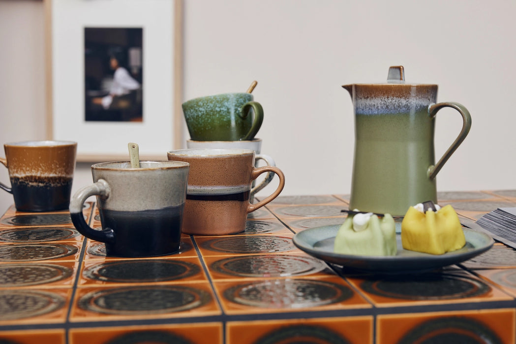 retro style green and brown stoneware teapot on an orange tile table next to cappuccino mugs and petit fours on a black dessert plate 