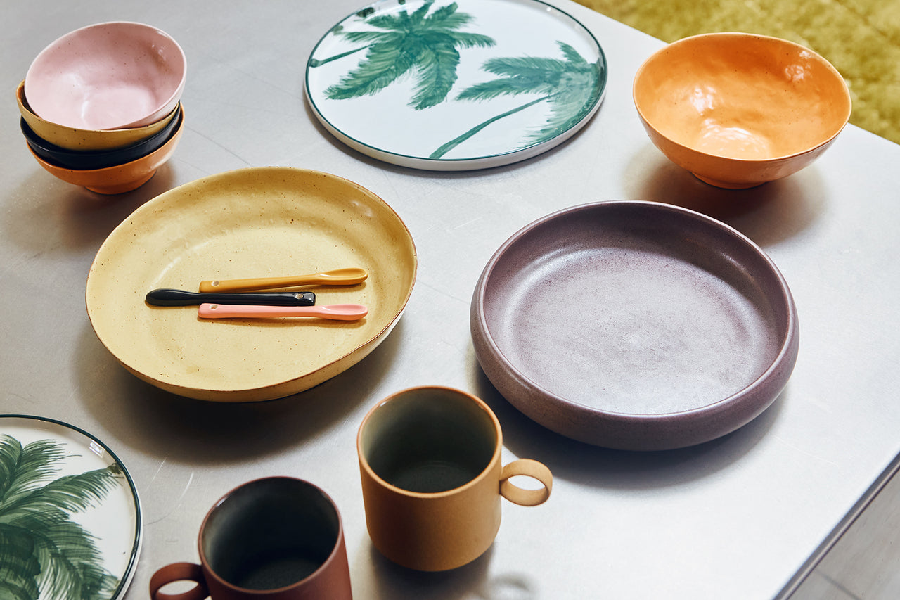 table with a deep porcelain yllow plate and spoons and a purple deep plate and a small plate with a green palm tree
