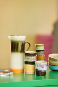 tall stoneware pitcher with orange, white and brown finish wth small coffee cups on a green table top