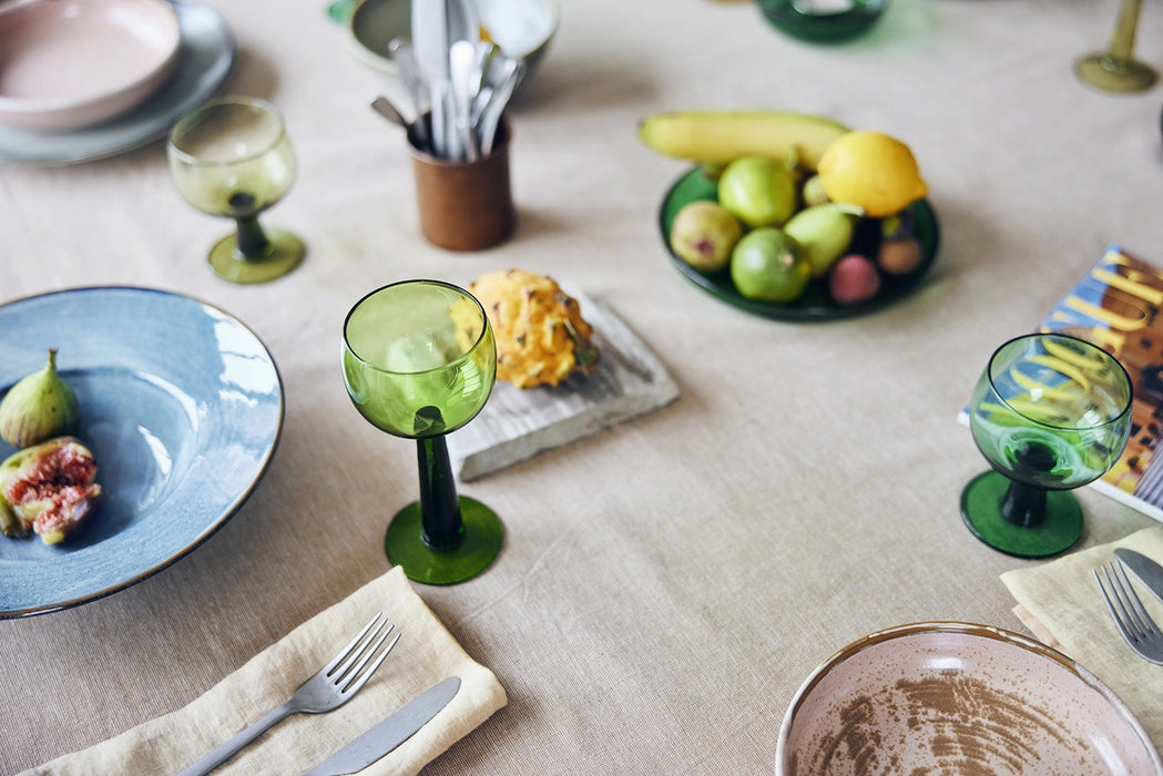 table with linen table cloth, green wine glasses blue and pink porcelain plates and a green plate made from glass filled with banana, lemon and limes