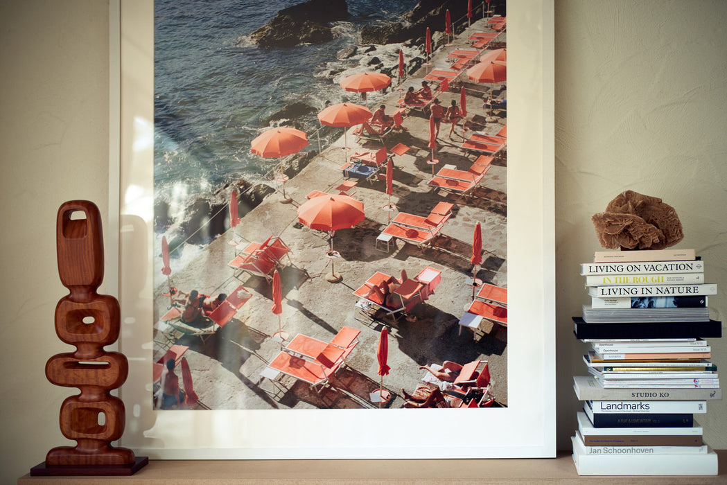 framed picture of orange parasols on the coast of Amalfi in a white frame next to a wooden sculpture and a stack of books