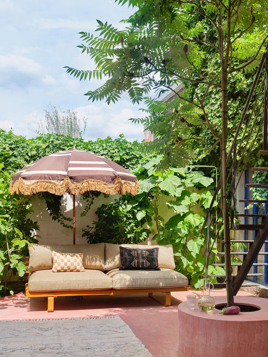 backyard patio with retro style parasol with fringes and an outdoor aluminium sofa with beige colored pillows against a wall with plants