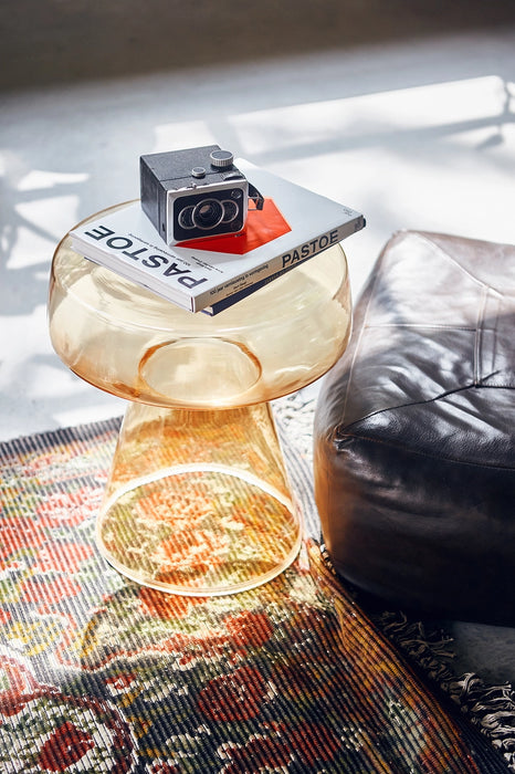 Amber glass colored Mid Century Modern style glass accent table  with a book and camera next to a leather pouf