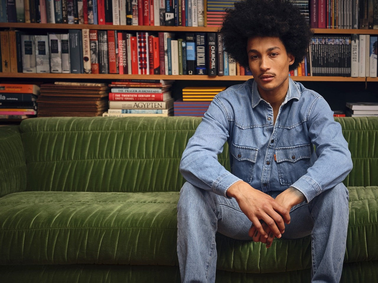 man with denim shirt sitting on the velvet green club couch in front of a bookshelf filled with books and striped storage boxes