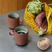 coral colored stoneware coffee mugs with ear on counter top with vegetables