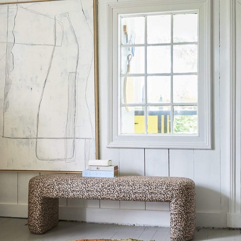 white painting next to a window with a floral print upholstered bench underneath with a stack of books