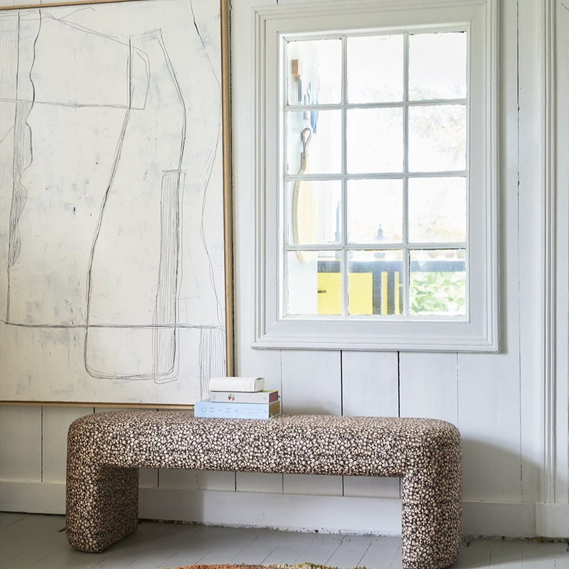 white painting next to a window with a floral print upholstered bench underneath with a stack of books