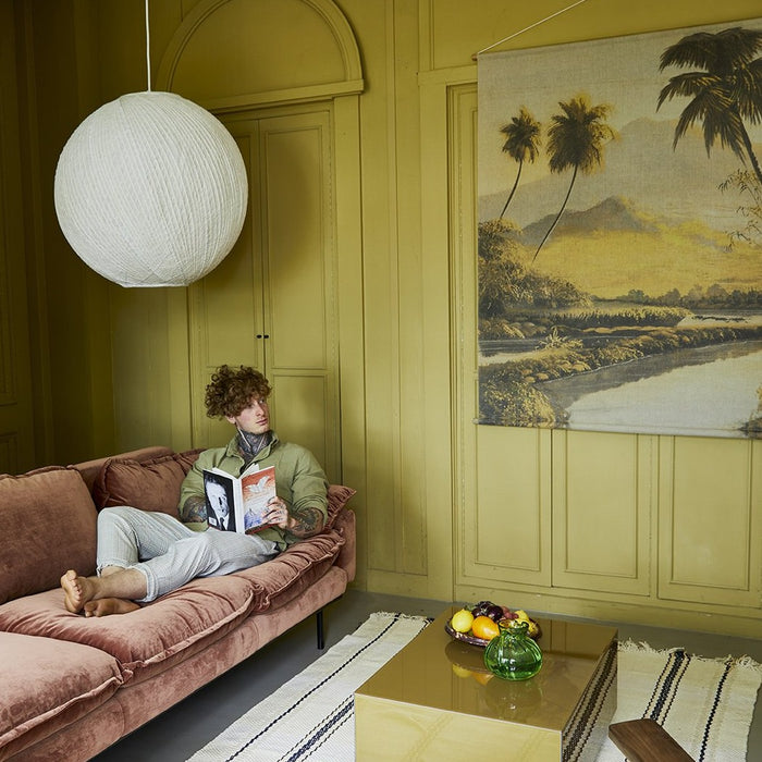 man reading a book on velvet sofa looking at a yellow colored jungle landscape wall mural