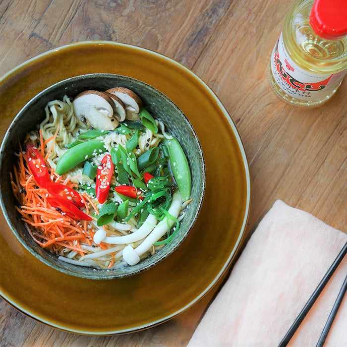 large brown dinner plate with on top a green colored ceramic noodle bowl with carrots, spring onion, mushrooms and red peppers with sesame seed and soy beans 
