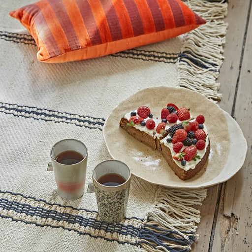 detail of fringes on a handwoven off white rug with black stripes and a red striped velvet pillow on top of it with two tea mugs made of stoneware and an organic shaped plated with carrot cake and strawberries